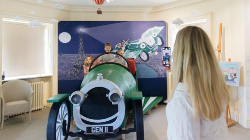 An adult taking a photo of three children sitting in a life-size model of an old car